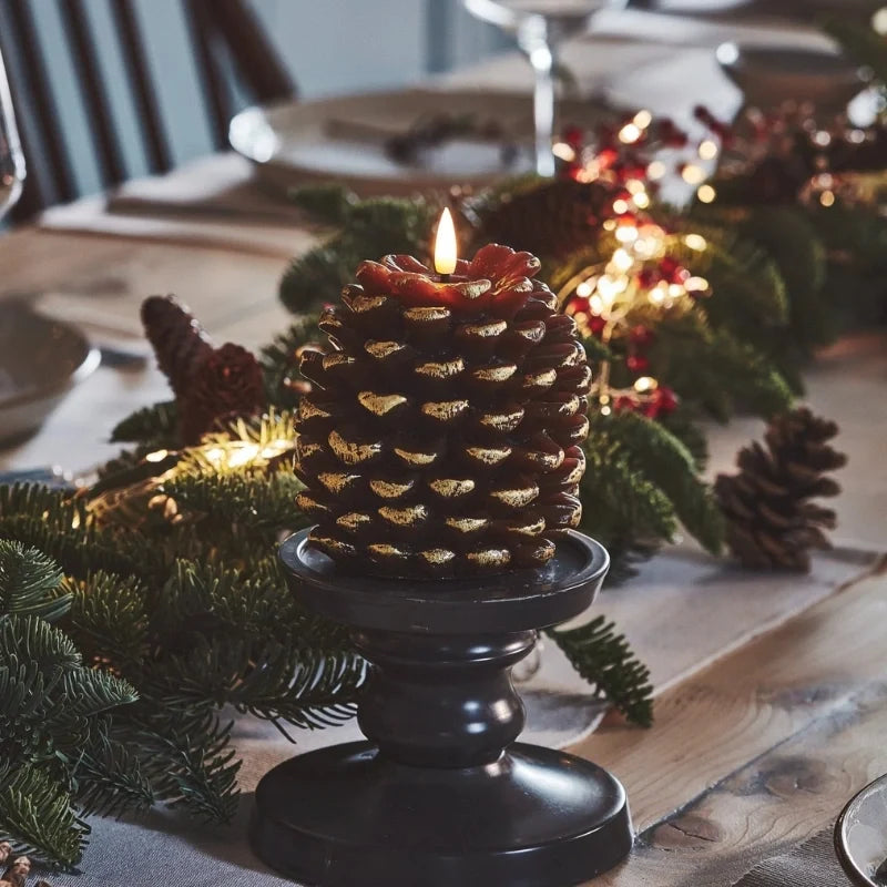 Golden brown pinecone candle on a black pedestal holder among greenery and string lights — Casa Norex