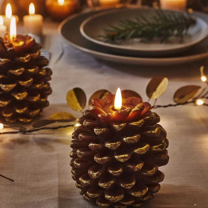 Golden brown pinecone candle with a lit LED flame on a dinner table set with plates and pine sprigs — Casa Norex