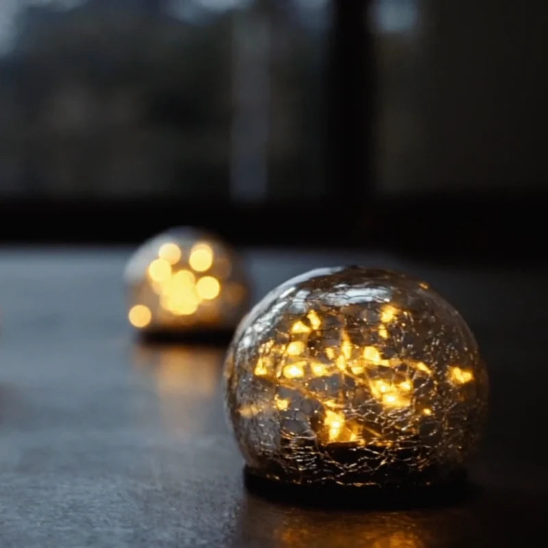 Cracked glass globe lit on a dark surface beside a smaller globe — Casa Norex